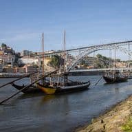 Traditionelle Boote auf dem Douro-Fluss mit der Dom-Luís-I-Brücke und Gebäuden am Hang in Porto, Portugal, unter einem klaren blauen Himmel.