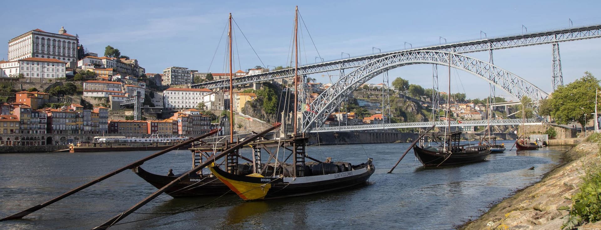 Traditionelle Boote auf dem Douro-Fluss mit der Dom-Luís-I-Brücke und Gebäuden am Hang in Porto, Portugal, unter einem klaren blauen Himmel.