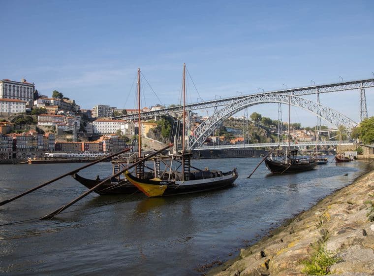 Traditionelle Boote auf dem Douro-Fluss mit der Dom-Luís-I-Brücke und Gebäuden am Hang in Porto, Portugal, unter einem klaren blauen Himmel.