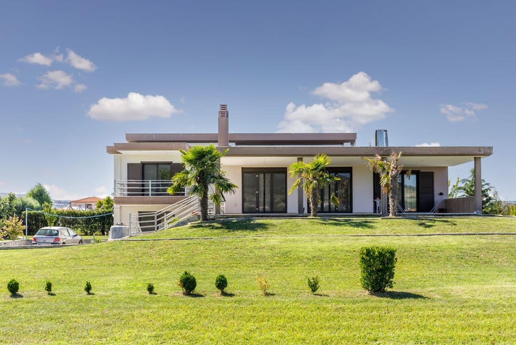 Modern single-story house with large windows, surrounded by a well-maintained lawn and small trees, under a clear blue sky.