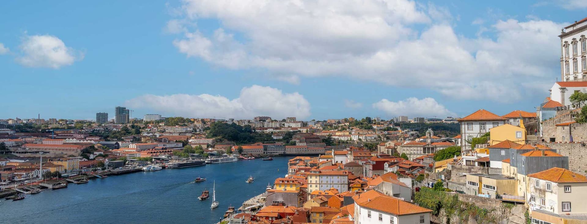 Scenic view of a riverside city with red-roofed buildings, a flowing river, boats, and a partly cloudy sky.