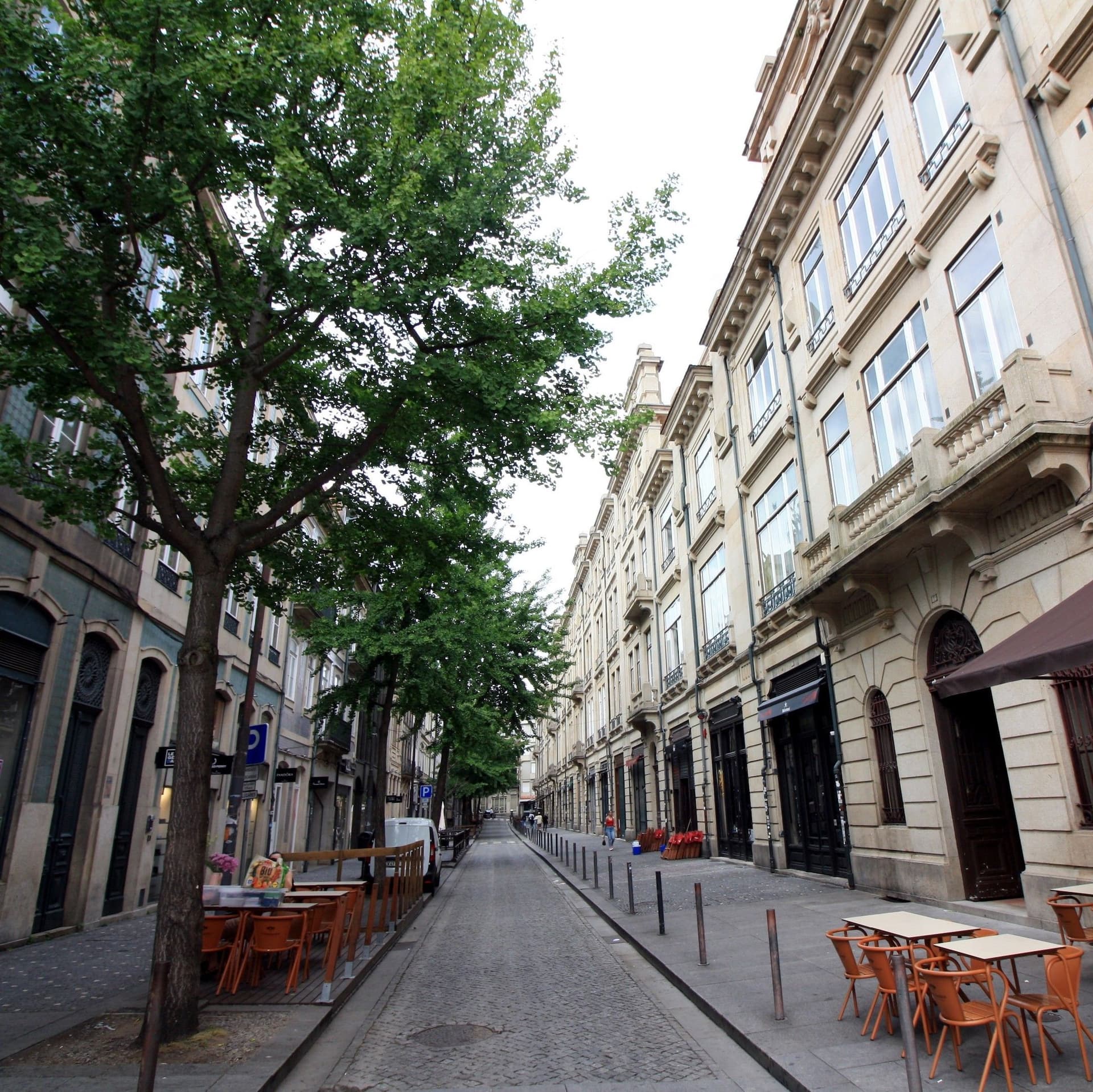 Narrow cobblestone street lined with historic buildings and outdoor café seating under trees on a clear day.