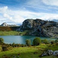 Vista panoràmica d'un llac blau envoltat de turons verds i muntanyes rocoses sota un cel ennuvolat.