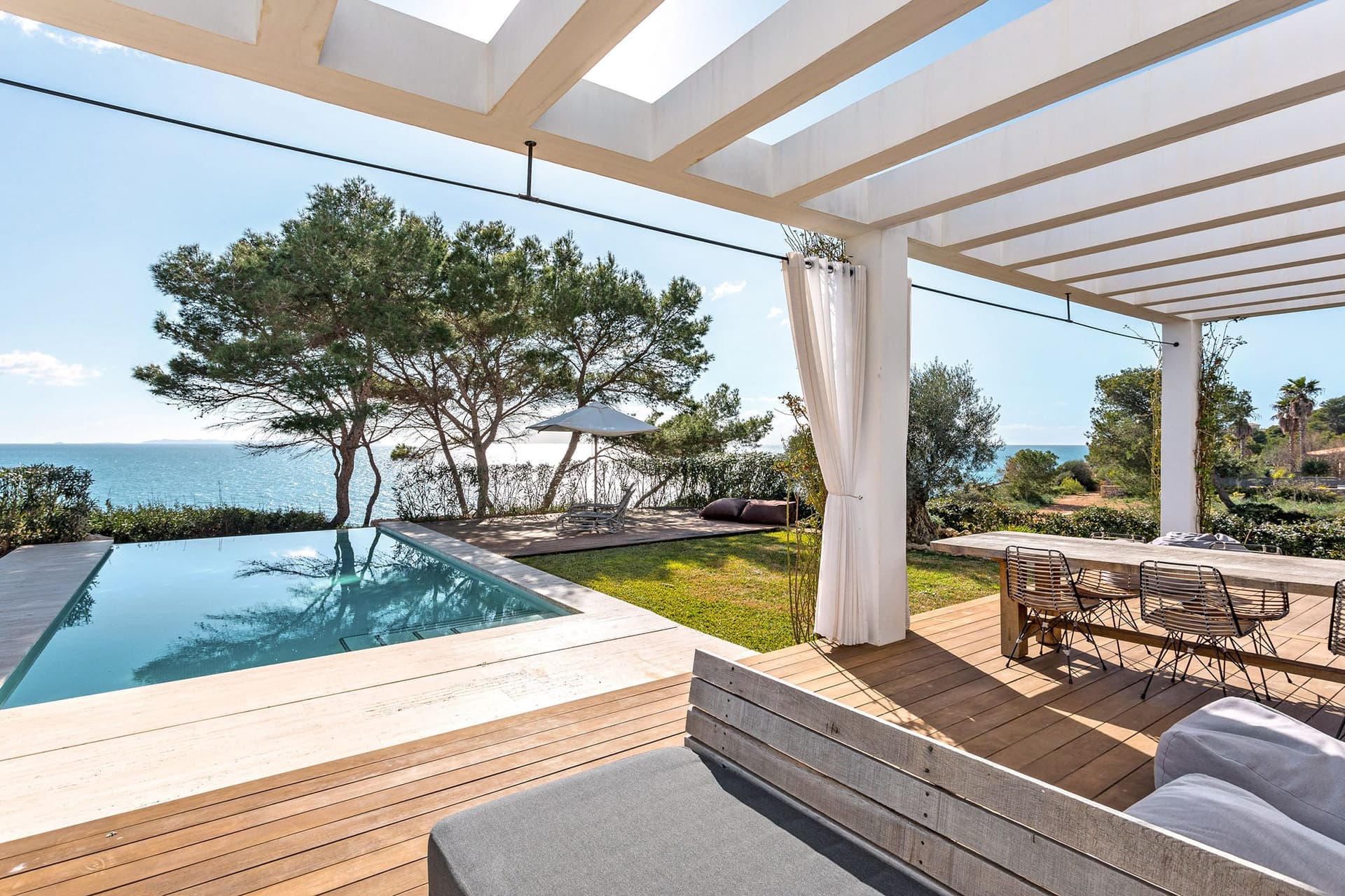 Moderne Villa auf Mallorca mit einem eleganten Außenbereich. Im Vordergrund befindet sich eine Holzterrasse mit einem gemütlichen Sitzbereich und einem langen Esstisch. Dahinter erstreckt sich ein Infinity-Pool, der nahtlos in die Aussicht auf das funkelnde Mittelmeer übergeht. Zwei große Bäume und ein Sonnenschirm bieten Schatten, während die klare, blaue Himmel und die üppige Vegetation die idyllische Szenerie vervollständigen. Dieses Bild vermittelt den Luxus und die Schönheit des Immobilienmarktes auf Mallorca.