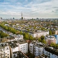 Luftaufnahme einer Stadtlandschaft mit dicht gedrängten Gebäuden, von Bäumen gesäumten Straßen und einem markanten Turm im Hintergrund unter einem teilweise bewölkten Himmel