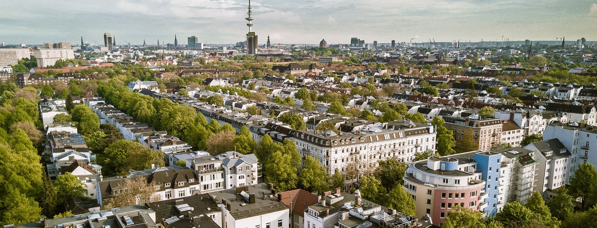 Luftaufnahme einer Stadtlandschaft mit dicht gedrängten Gebäuden, von Bäumen gesäumten Straßen und einem markanten Turm im Hintergrund unter einem teilweise bewölkten Himmel