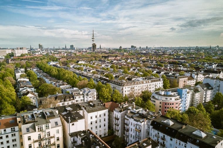 Luftaufnahme einer Stadtlandschaft mit dicht gedrängten Gebäuden, von Bäumen gesäumten Straßen und einem markanten Turm im Hintergrund unter einem teilweise bewölkten Himmel