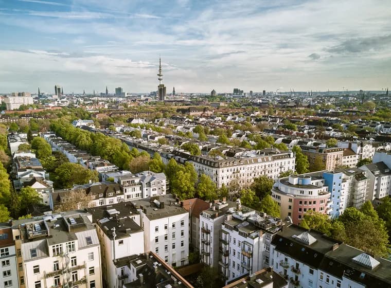 Luftaufnahme einer Stadtlandschaft mit dicht gedrängten Gebäuden, von Bäumen gesäumten Straßen und einem markanten Turm im Hintergrund unter einem teilweise bewölkten Himmel