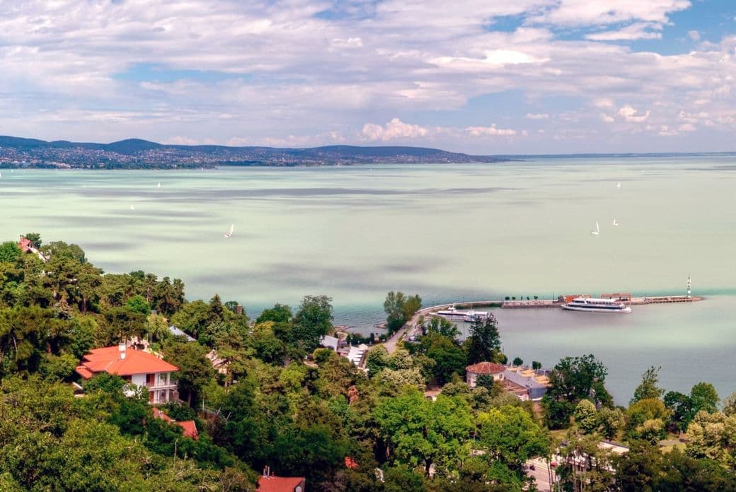 A scenic view of hee lakeside Balaton, Hungary, with red-roofed houses, lush greenery, a pier, and sailboats on a calm, expansive lake under a partly cloudy sky.