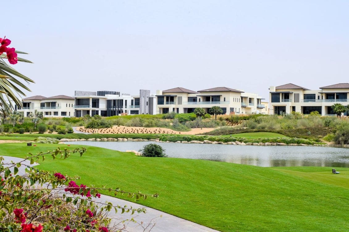 Luxurious houses near a serene lake, surrounded by lush green lawns and vibrant flowers under a clear blue sky.