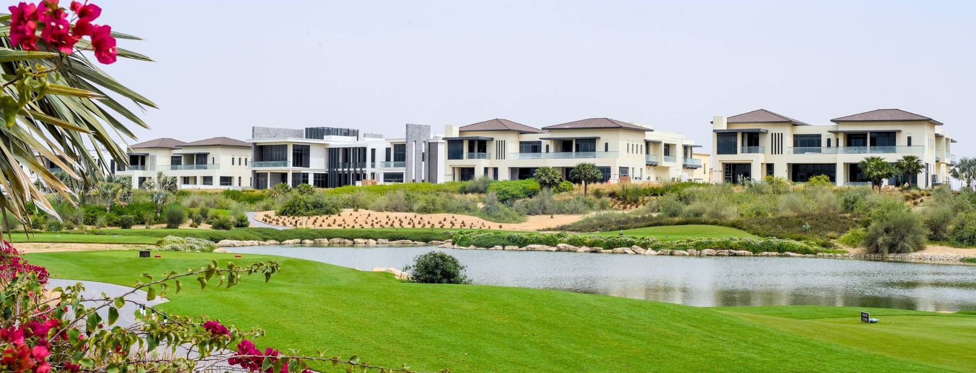 Luxurious houses by a serene lake with vibrant green lawns and blooming flowers under a clear blue sky.