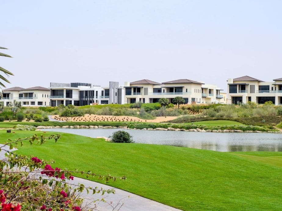 Luxurious houses by a serene lake with vibrant green lawns and blooming flowers under a clear blue sky.