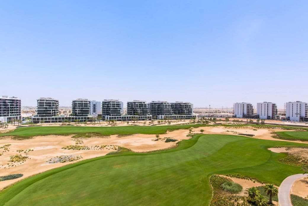 Eine Weitwinkelaufnahme von einem grünen Golfplatz mit modernen Apartmentgebäuden im Hintergrund unter einem klaren blauen Himmel.