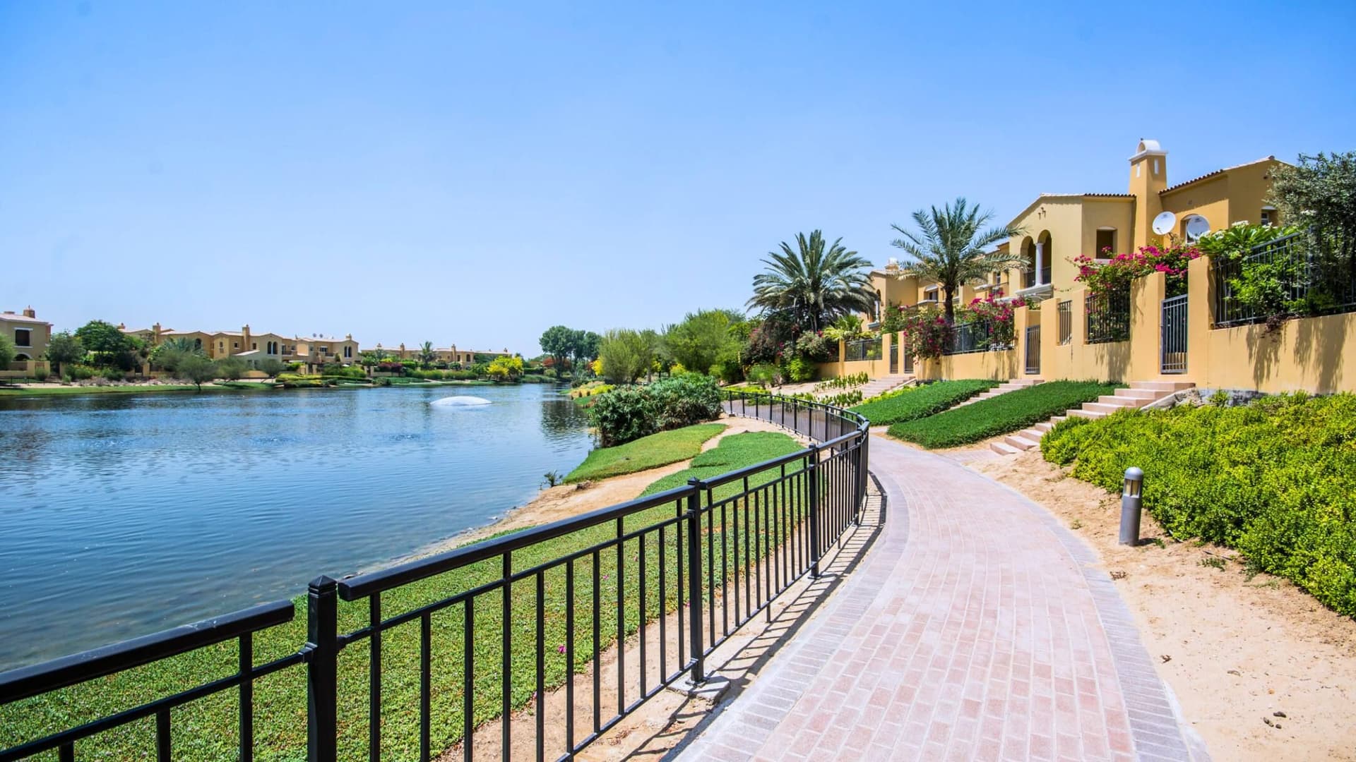 Lakeside path in Arabian Ranches with black railing, flanked by grass and palm trees, leads to beige villas under a clear blue sky.