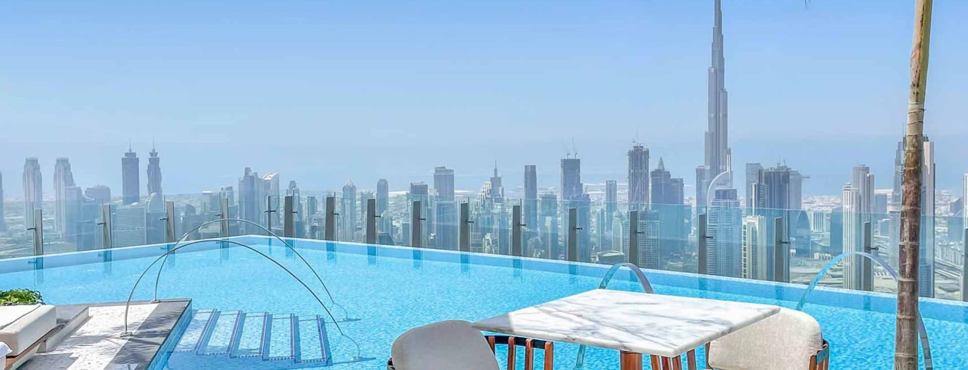 Rooftop infinity pool with a view of a city skyline and a tall tower, featuring a seating area with chairs and a table under clear blue skies.