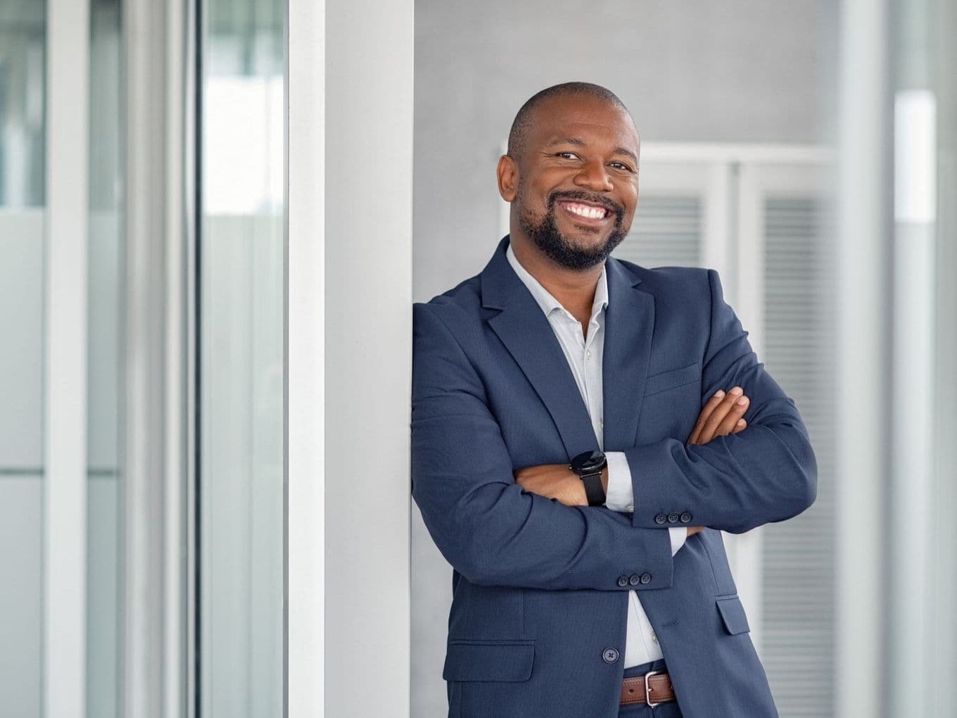 Professional businessman in a navy suit smiling confidently with arms crossed in a modern office setting