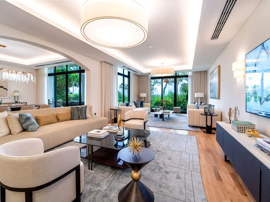 Luxurious living room with modern furniture, large windows, elegant lighting, and a mix of neutral tones. Greenery visible outside.