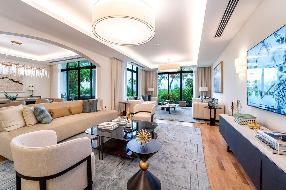 Helles, modernes Wohnzimmer mit beigen Sofas, Stühlen und einem grauen Teppich. Große Fenster bieten einen Blick auf üppiges Grün. Ein Fernseher hängt an der Wand.