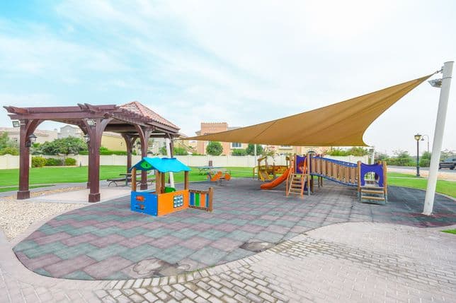 Outdoor kids’ play area with shaded climbing frames, slides, swings, and a pergola set in green open space.