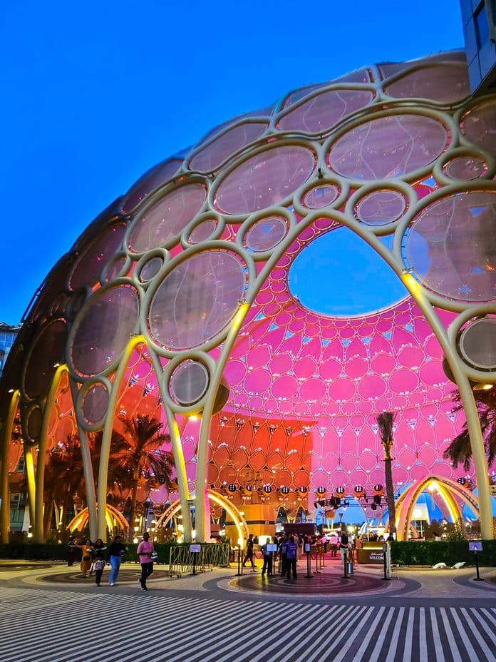 A futuristic dome with circular patterns, illuminated in pink and purple hues, with people and palm trees nearby under a twilight sky.