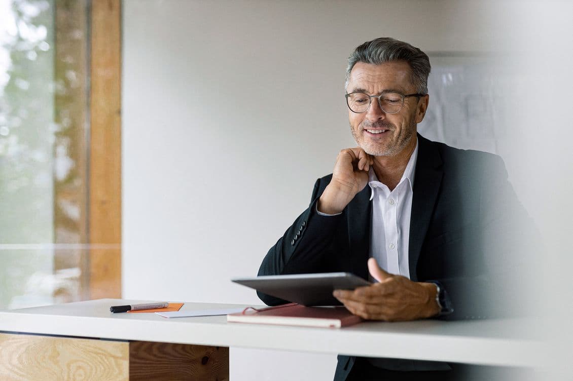 Smiling man with glasses in a suit, sitting at a desk, holding a tablet, and looking engaged, with papers and a pen nearby.