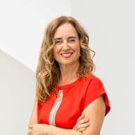 Smiling woman with long curly hair in a red dress stands against a white abstract background.