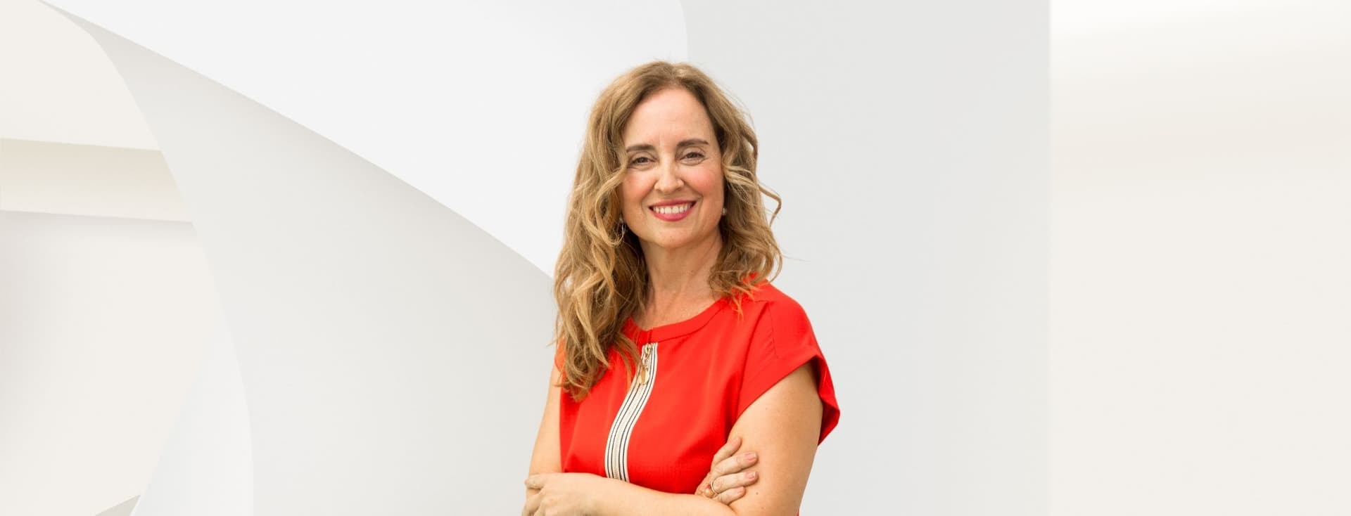 Smiling woman with long curly hair in a red dress stands against a white abstract background.