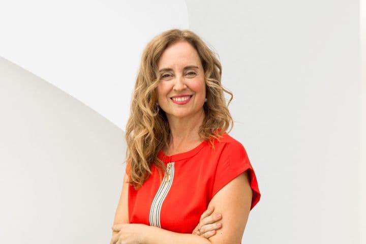 Smiling woman with long curly hair in a red dress stands against a white abstract background.