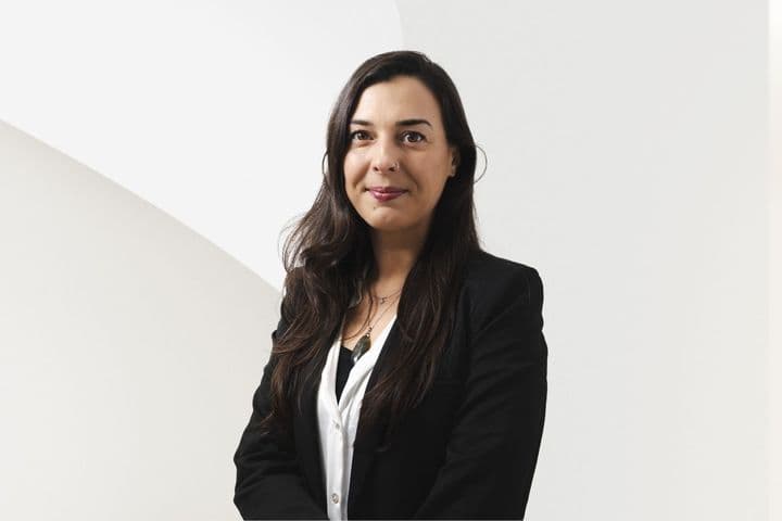 Woman in a black blazer and white shirt smiling against a minimalistic white background.