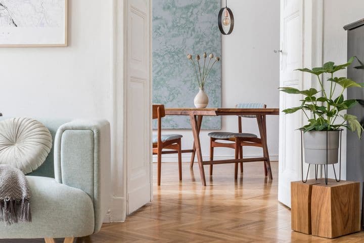 Heller, offener Wohnbereich im skandinavischen Stil mit einem pastellgrünen Loungesessel mit dekorativen Kissen, abstrakter Wandkunst, einem schwarzen Vintage-Klavier, hölzernen Esszimmermöbeln und Fischgrätparkettboden, der eine perfekte Mischung aus modernen und klassischen Elementen darstellt