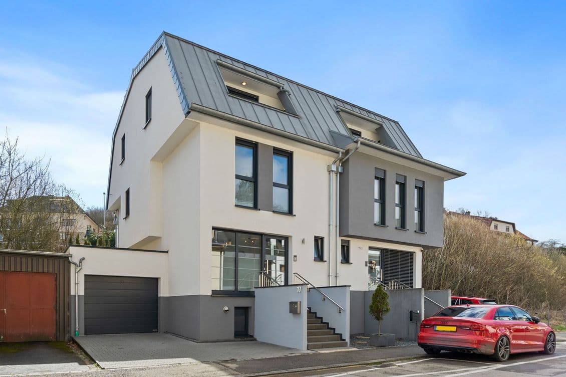 Modern two-story house with a gray roof, large windows, and an attached garage. A red car is parked on the street in front.