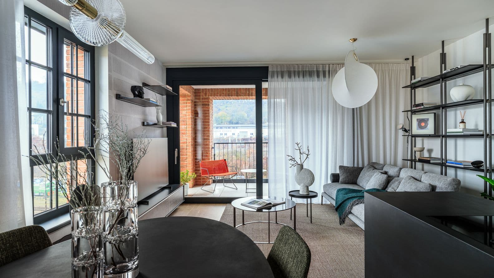 Moderní obývací pokoj s šedou pohovkou, černými policemi a kulatým jídelním stolem, za skleněnými dveřmi navazuje lodžie s výhledem ven.