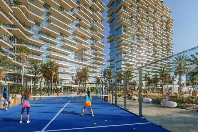 People playing tennis on a blue court surrounded by palm trees, with tall, modern buildings in the background.