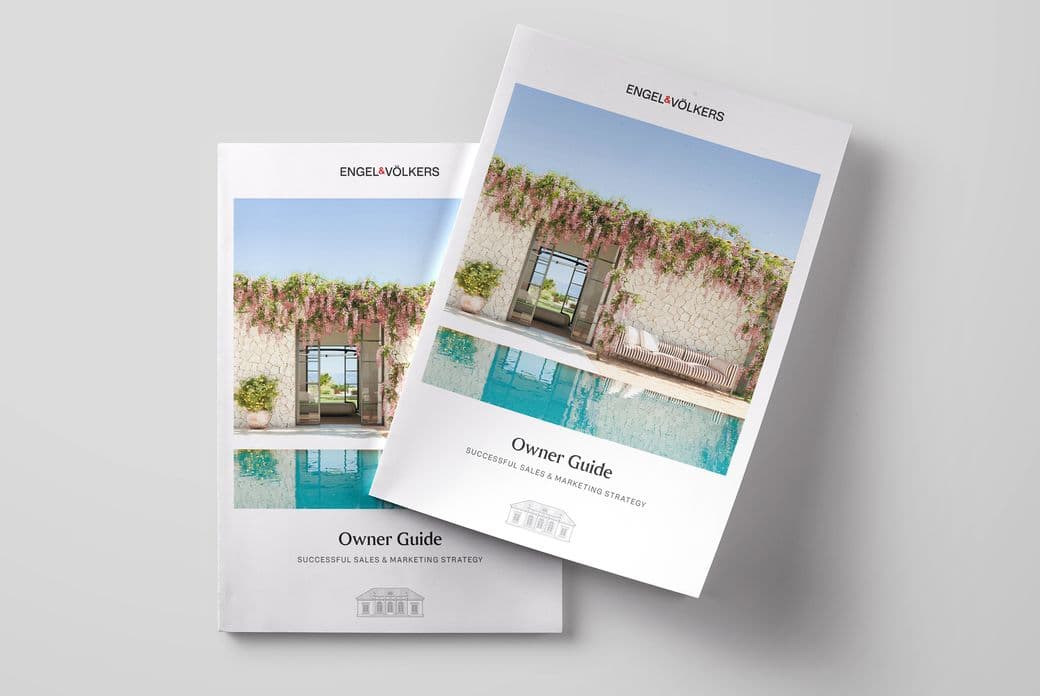 Two copies of the Engel & Völkers 'Owner Guide: Successful Sales & Marketing Strategy' brochure placed on a light grey surface. The cover features a luxurious poolside view with a stone wall, open doorway, flowering vines, and stylish outdoor furniture.