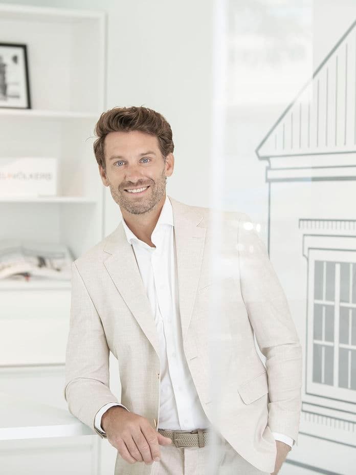 Smiling man in a light beige suit leaning on a white counter in a bright, modern office with framed photos on shelves.