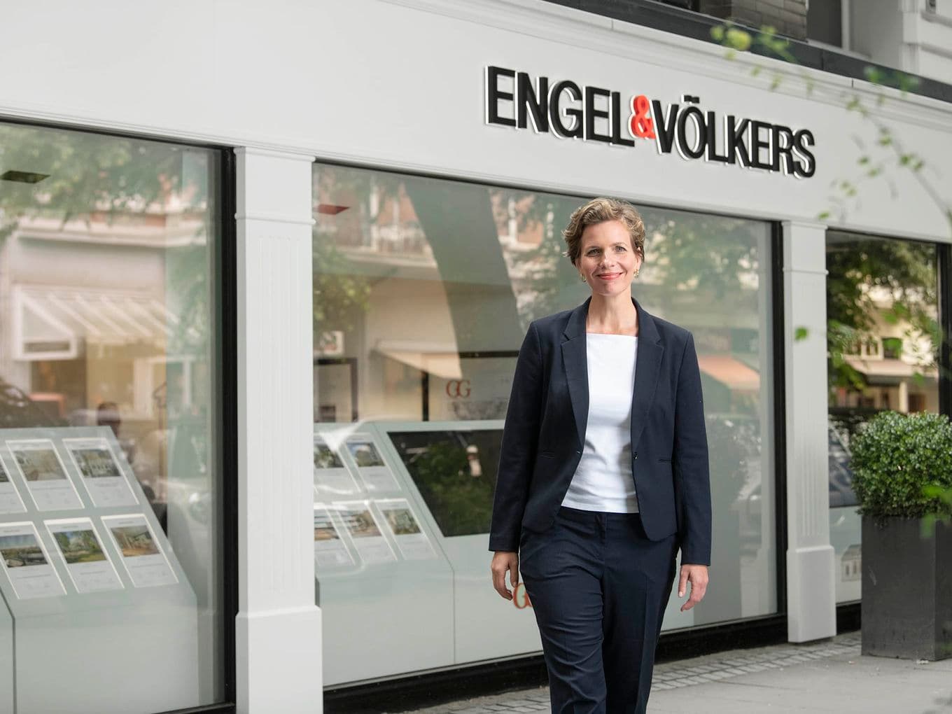 Smiling woman in a navy blazer and white top stands outside Engel & Völkers real estate storefront with property brochures in display windows.