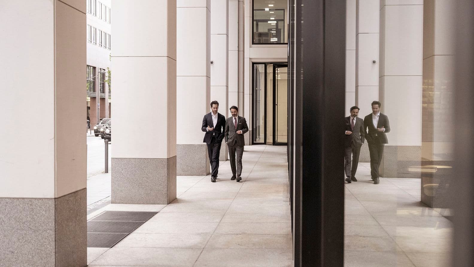 Zwei Männer in Anzügen gehen einen Korridor zwischen hohen Säulen entlang, wobei sich ihr Spiegelbild an einer Glaswand abzeichnet. Urbane Umgebung mit moderner Architektur.