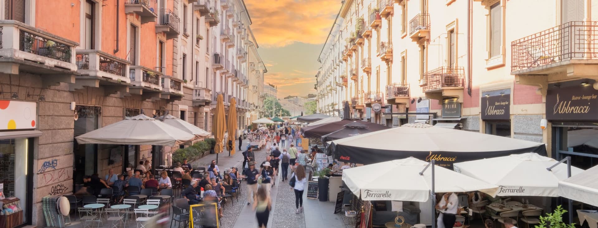 A bustling European street with outdoor cafes, people walking, and a vibrant sunset sky.