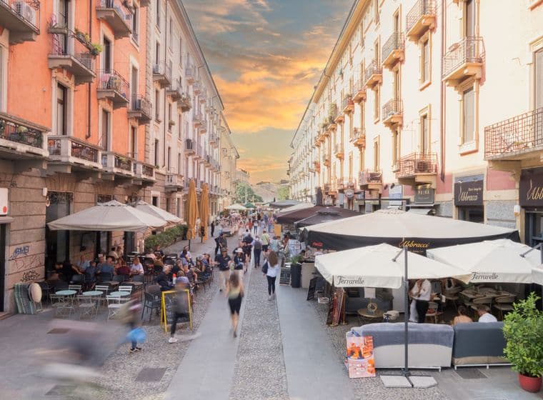 A bustling European street with outdoor cafes, people walking, and a vibrant sunset sky.