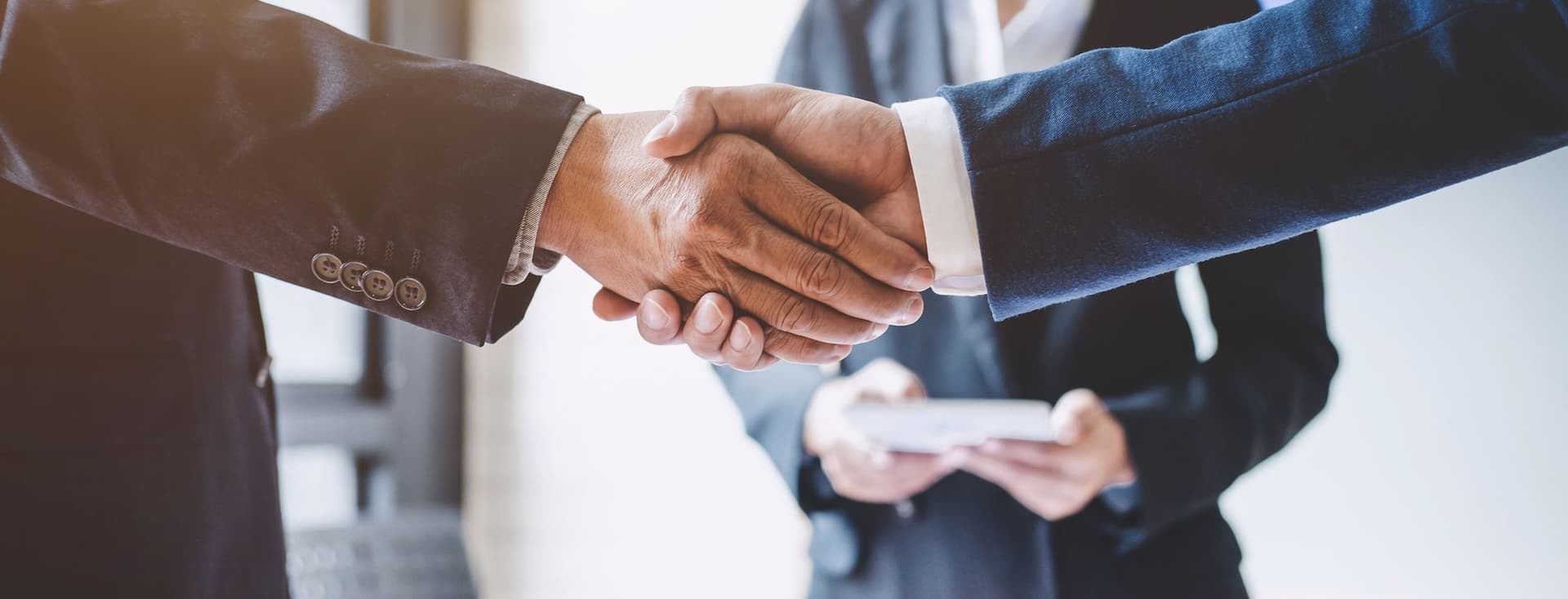 Two people in business suits shaking hands over a table with documents, while another person stands in the background holding a tablet.
