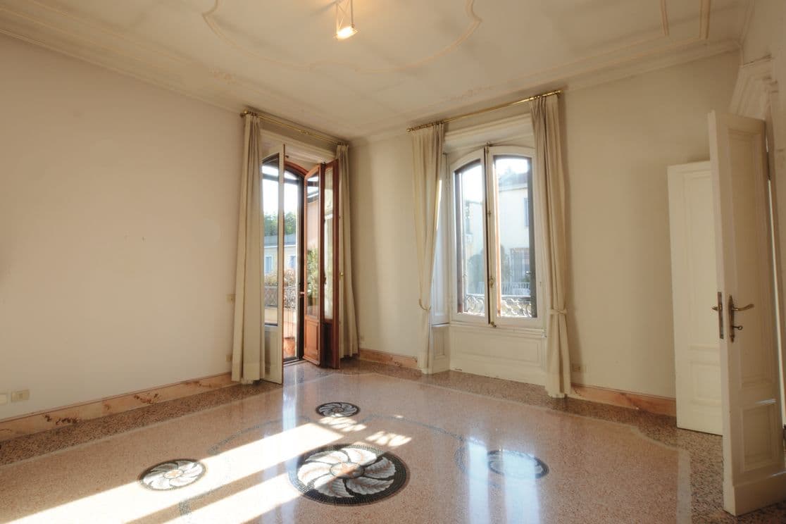 Heller, leerer Raum mit verzierter Decke, großen Fenstern und offenen Türen, die auf einen Balkon führen. Sonnenlicht wirft Muster auf den polierten Boden.