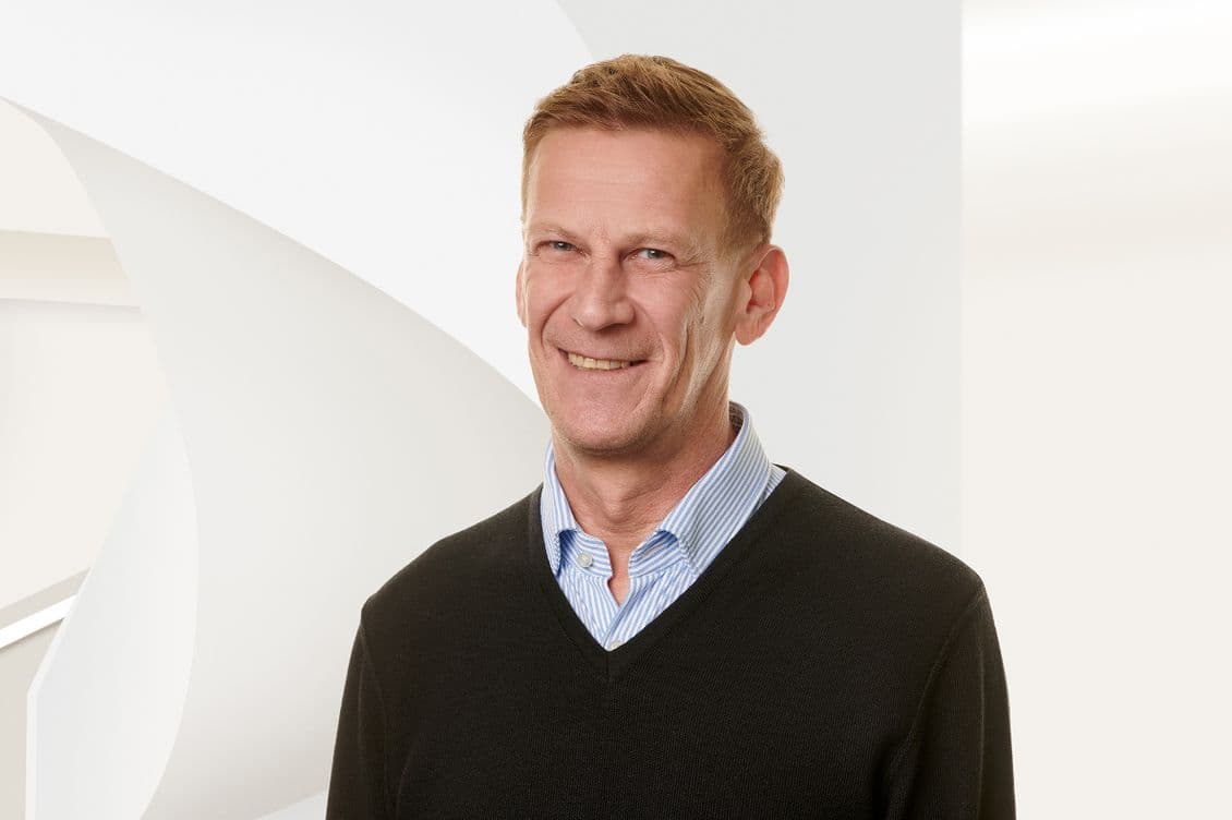 Smiling man in a black sweater over a blue striped shirt, standing against a light, abstract background.