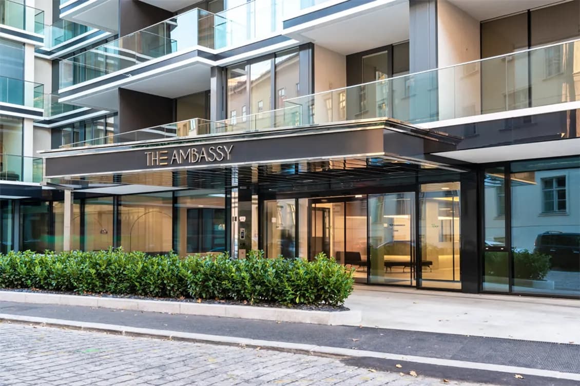 Moderner Gebäudeeingang mit großen Glastüren und Fenstern, beschriftet mit „The Embassy“. Umgeben von Grünflächen und einer Kopfsteinpflasterauffahrt.