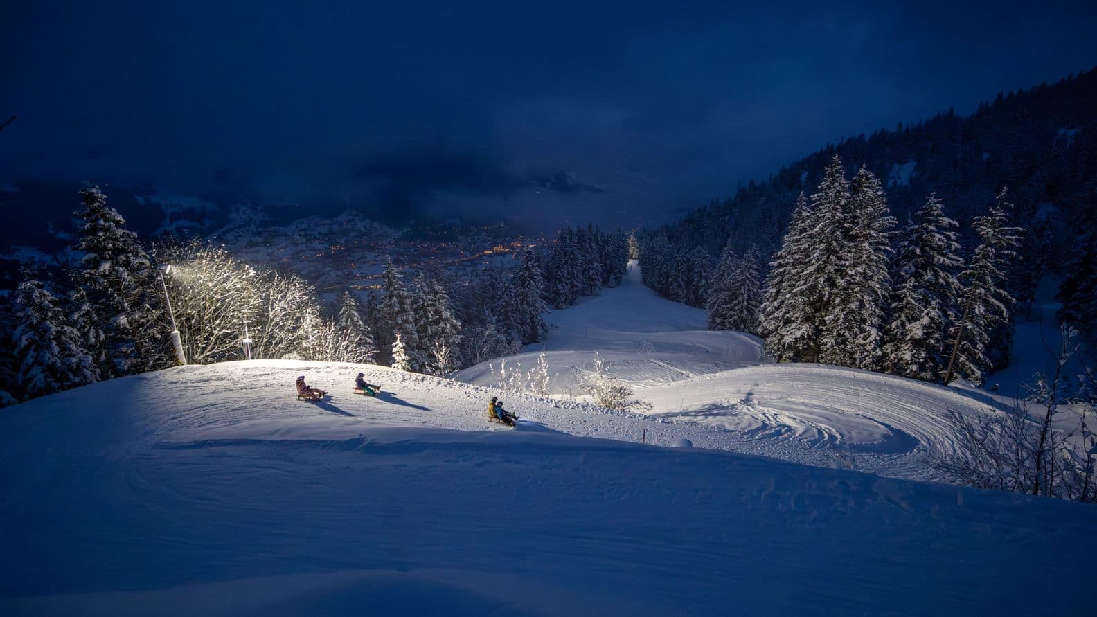Skiers glide down a snowy, illuminated slope at night, surrounded by snow-covered trees, with a village visible in the background.