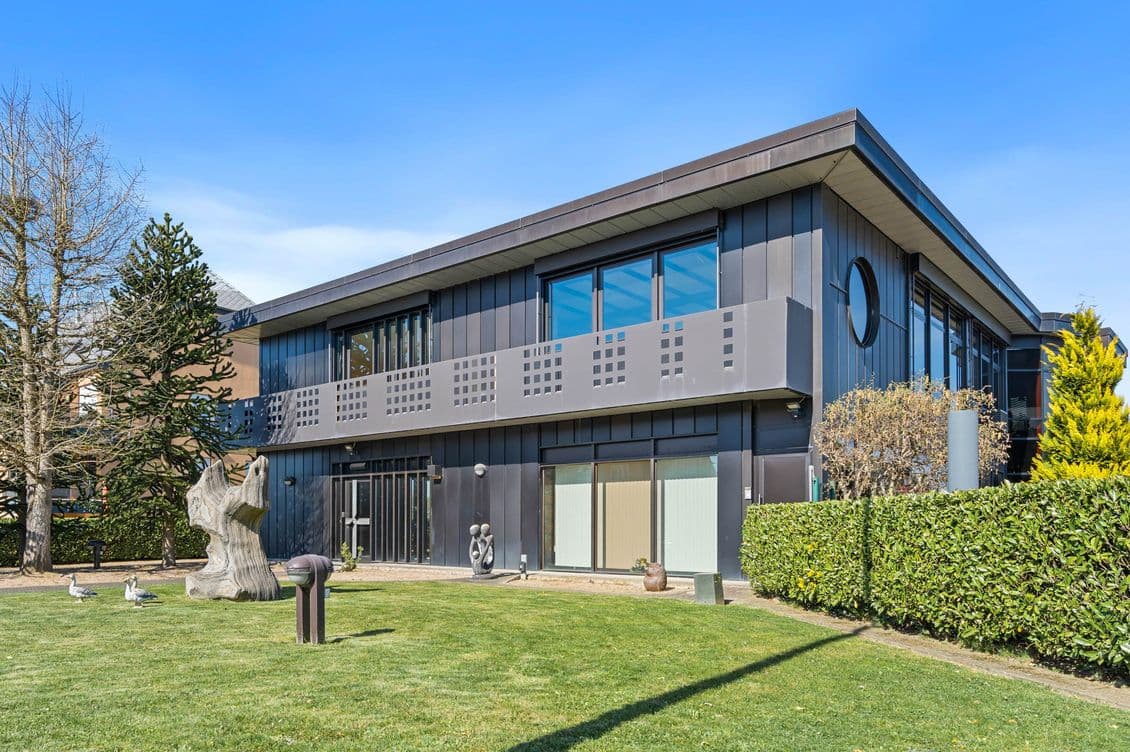 Modern two-story building with large windows, geometric facade, and a manicured lawn featuring sculptures and trimmed hedges. Blue sky above.