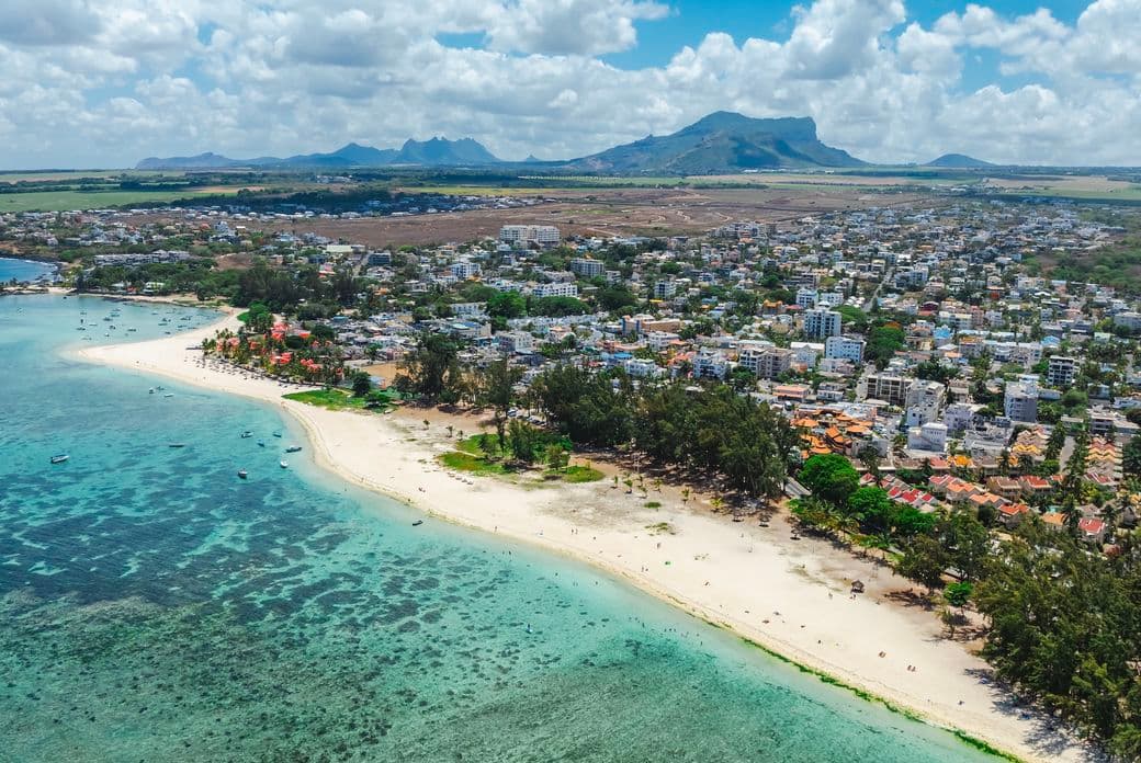 Aerial view of a coastal area with a sandy beach, turquoise waters, and lush greenery, set against a backdrop of mountains and scattered clouds.