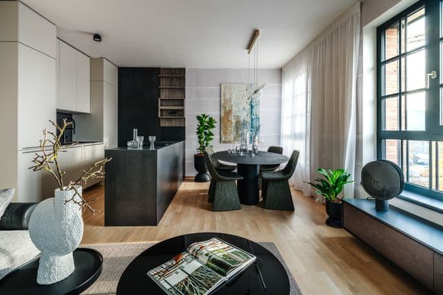 Moderní kuchyně s ostrůvkem a jídelním stolem u velkého okna, interiér doplňují rostliny, dekorace a obraz na stěně.