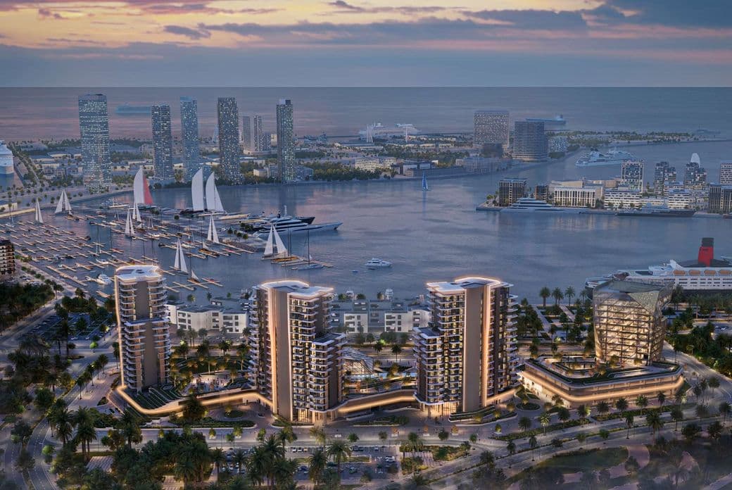 Aerial view of a modern marina with sailboats, yachts, and illuminated skyscrapers against a sunset backdrop, surrounded by lush greenery.