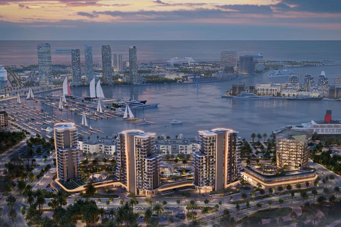 Aerial view of a modern marina with sailboats, yachts, and illuminated skyscrapers against a sunset backdrop, surrounded by lush greenery.