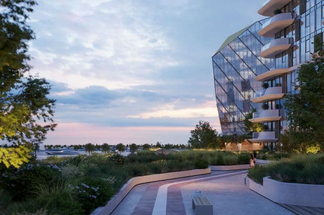 Modern building with curved balconies beside a landscaped promenade, overlooking a scenic sunset with scattered clouds and palm trees.